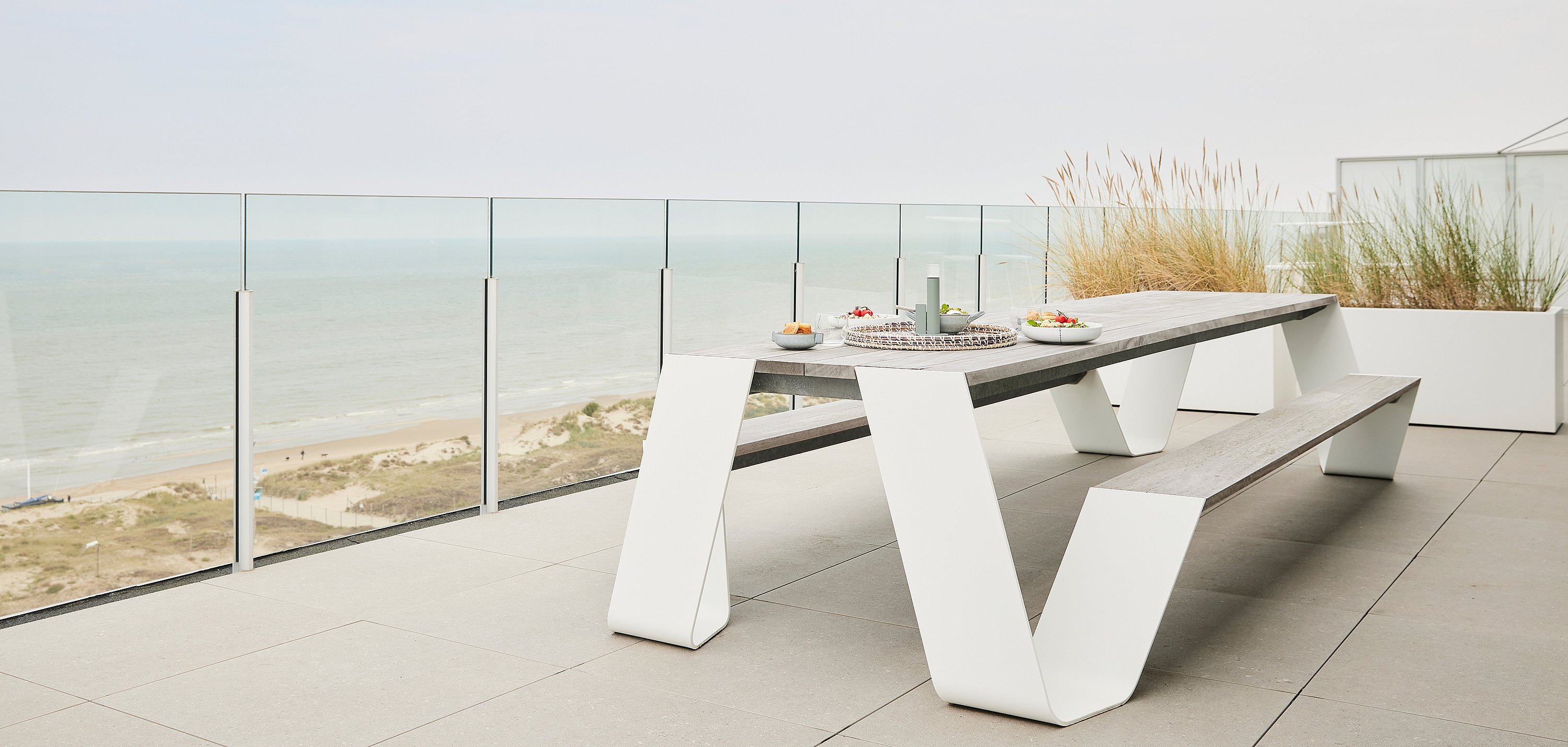 Lege Hopper-picknicktafel op een dakterras met uitzicht op de kustlijn, met duingrassen in plantenbakken achter de tafel.