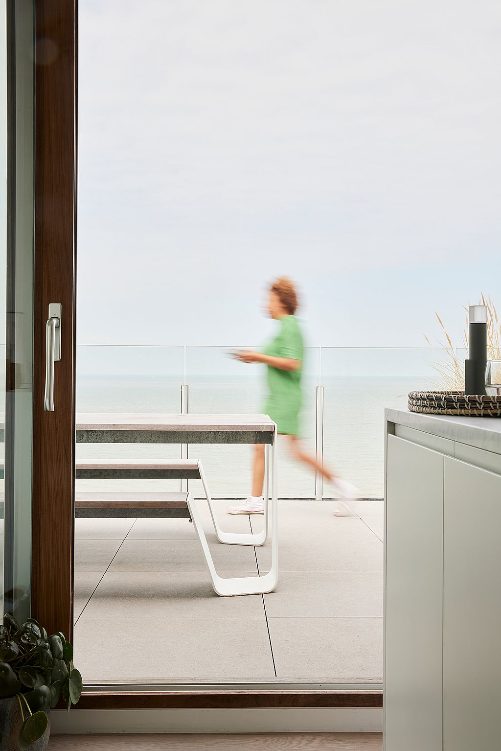 Zijaanzicht van een witte Hopper-picknicktafel gezien door een open deuropening, met een wazig persoon die voorbij loopt en de zee voorbij de glazen balustrade.