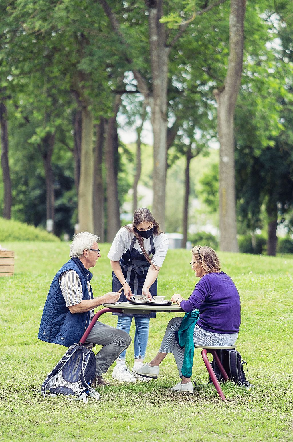 Eine Bedienung stellt Getränke und Speisen auf einen Virus-Tisch für zwei ältere Besucher, die auf der Rasenfläche im Park sitzen.