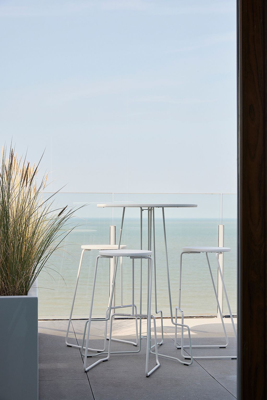 Witte Tiki hoge tafel met bijpassende krukken naast een glazen reling, met uitzicht op duinen, daken en de verre kust.