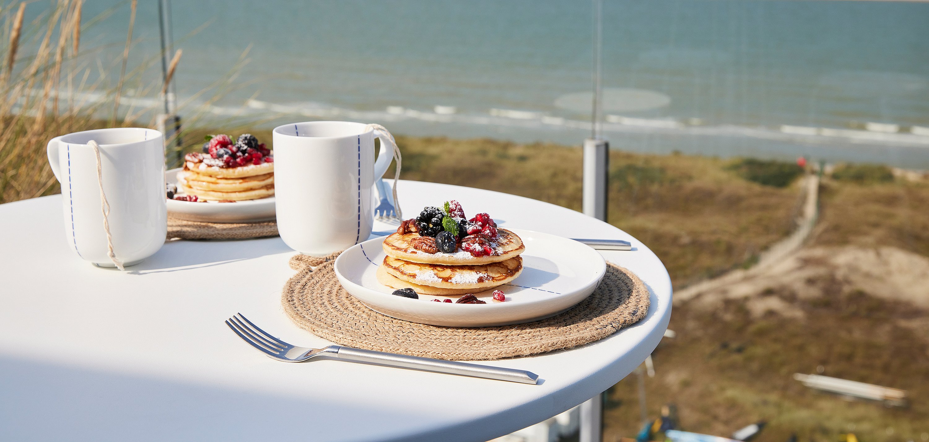 Ontbijtdetail met pannenkoeken, bessen, mokken en bestek op een ronde witte tafel met uitzicht op het strand en de zee beneden.