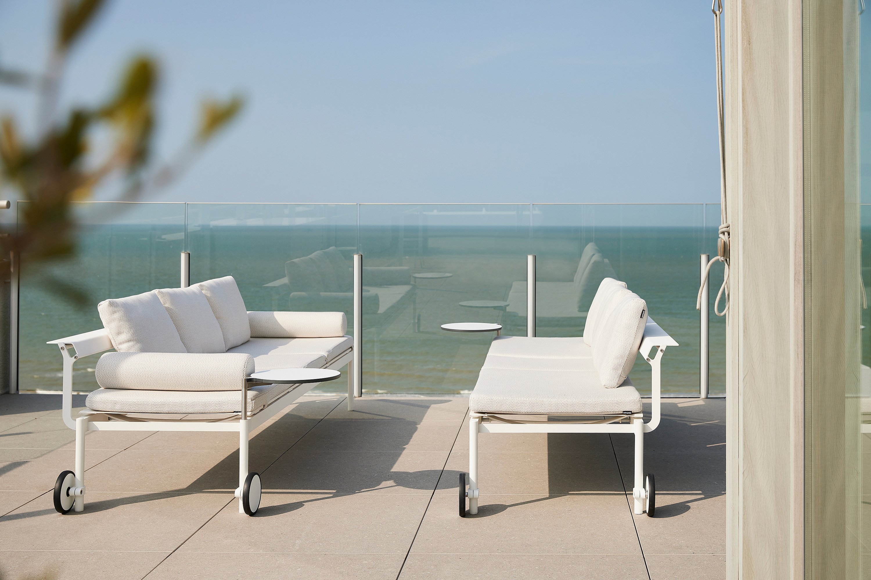Twee witte Sol + Luna lounge banken staan tegenover elkaar op een terras met kleine bijzettafels, tegen een glazen reling en open uitzicht op zee.