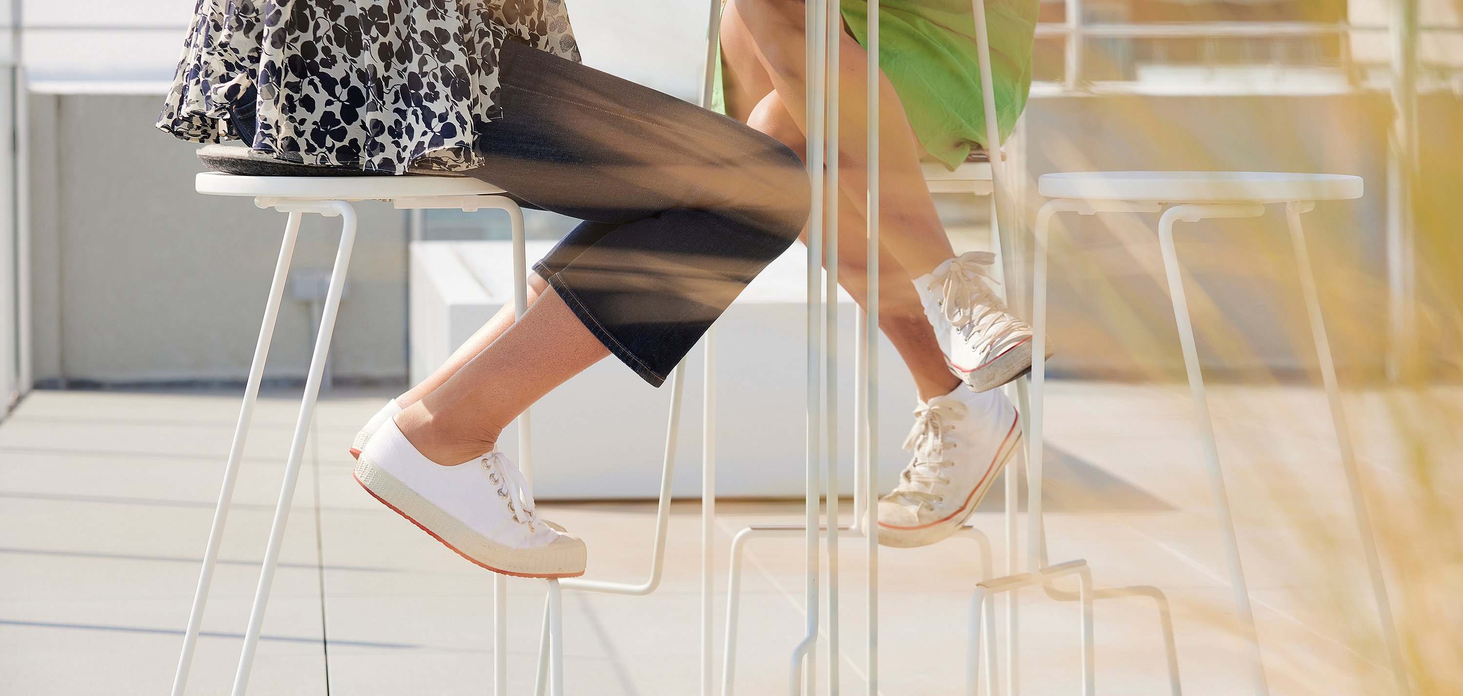 Close-up van twee personen zittend aan een hoge buitentafel, met witte sneakers die rusten op de krukvoeten in warm zonlicht.