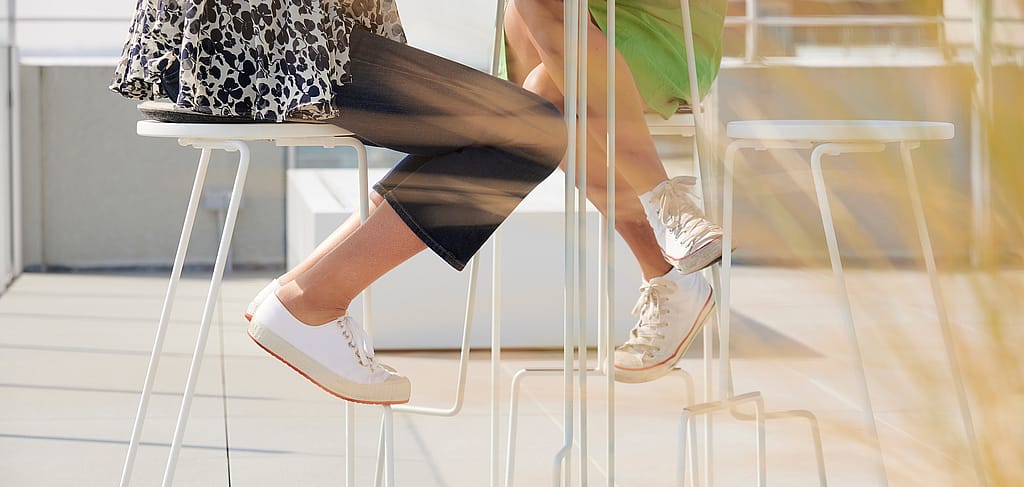 Close-up van twee personen zittend aan een hoge buitentafel, met witte sneakers die rusten op de krukvoeten in warm zonlicht.