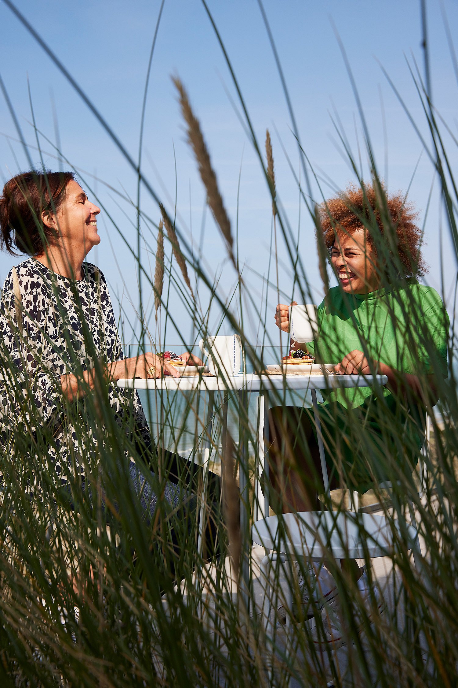 Twee vrouwen lachen samen tijdens het ontbijt aan een witte terrastafel, gedeeltelijk omlijst door hoge duingrassen tegen een blauwe lucht.