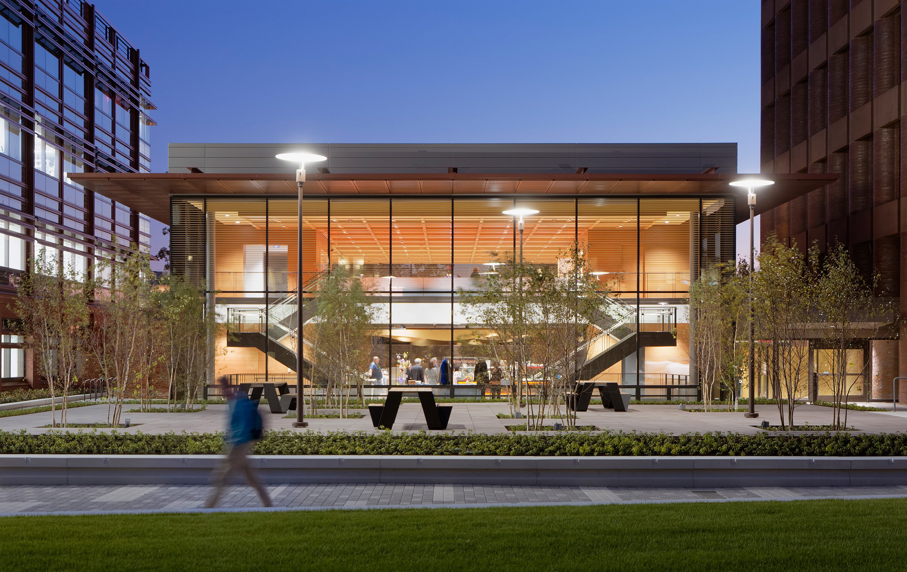 Der gläserne Pavillon des Yale Science Building erstrahlt in der Abenddämmerung warm zwischen zwei höheren Gebäuden. Schwarze Hopper-Picknicktische laden auf dem Vorplatz zum Verweilen ein, im Vordergrund ist ein Fußgänger verschwommen zu erkennen.