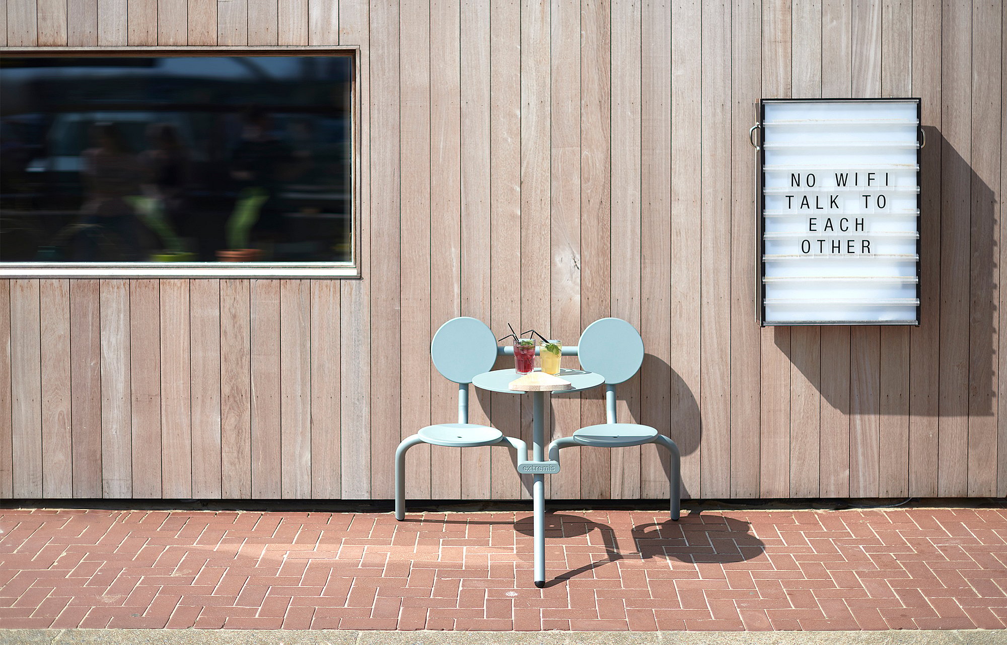 Pale blue Bistroo 2-seater set against a wooden restaurant façade, with two colorful drinks on the table and a lightbox sign reading ‘No wifi, talk to each other.