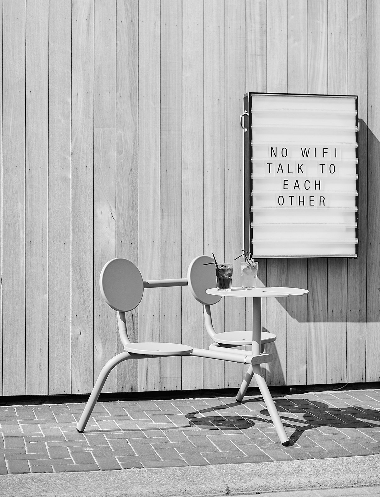 Black-and-white photo of a Bistroo 2-seater with two drinks on the table, positioned beside a lightbox sign against vertical wooden cladding.