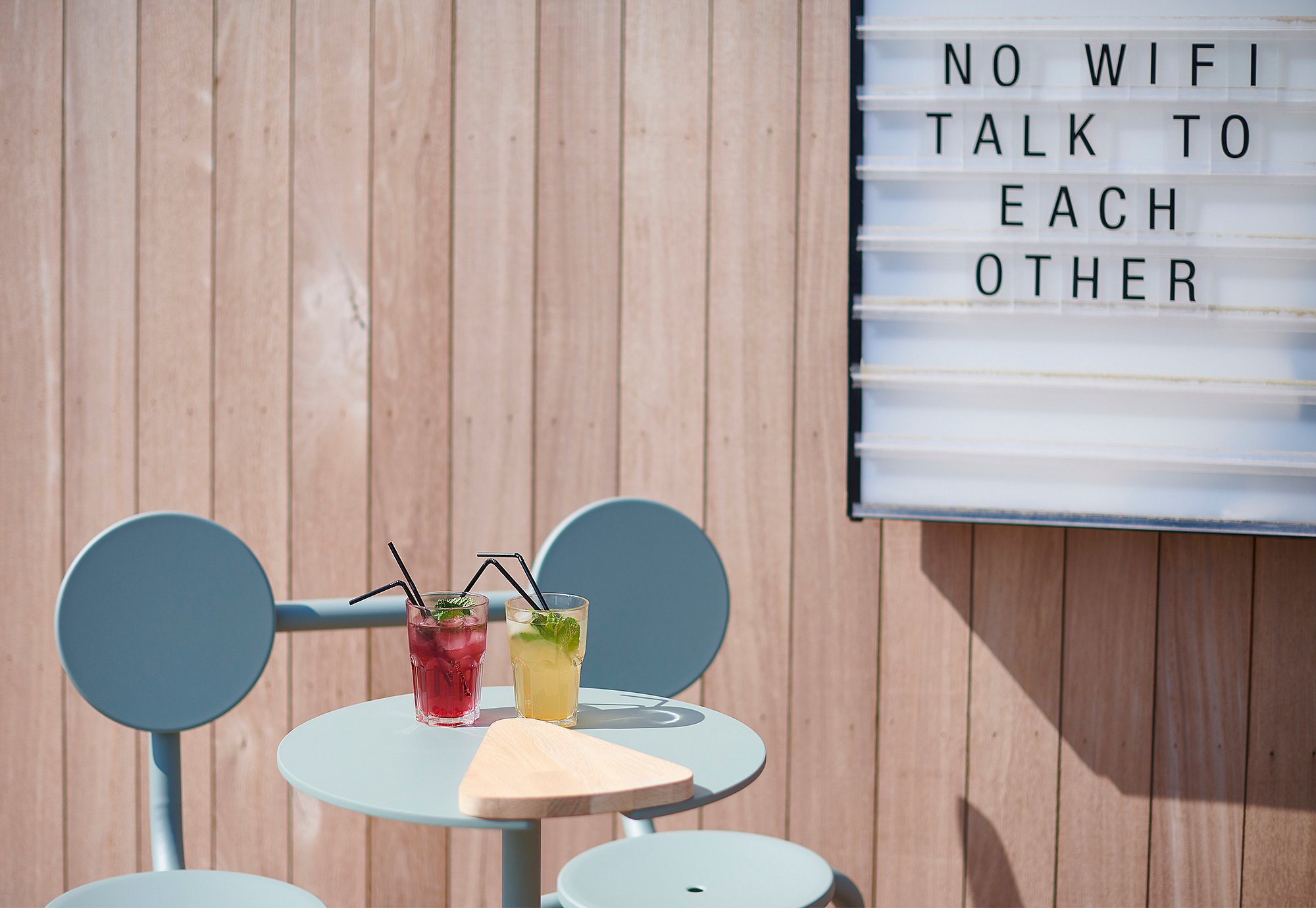 Pale blue Bistroo 2-seater set against a wooden restaurant façade, with two colorful drinks on the table and a lightbox sign reading ‘No wifi, talk to each other.