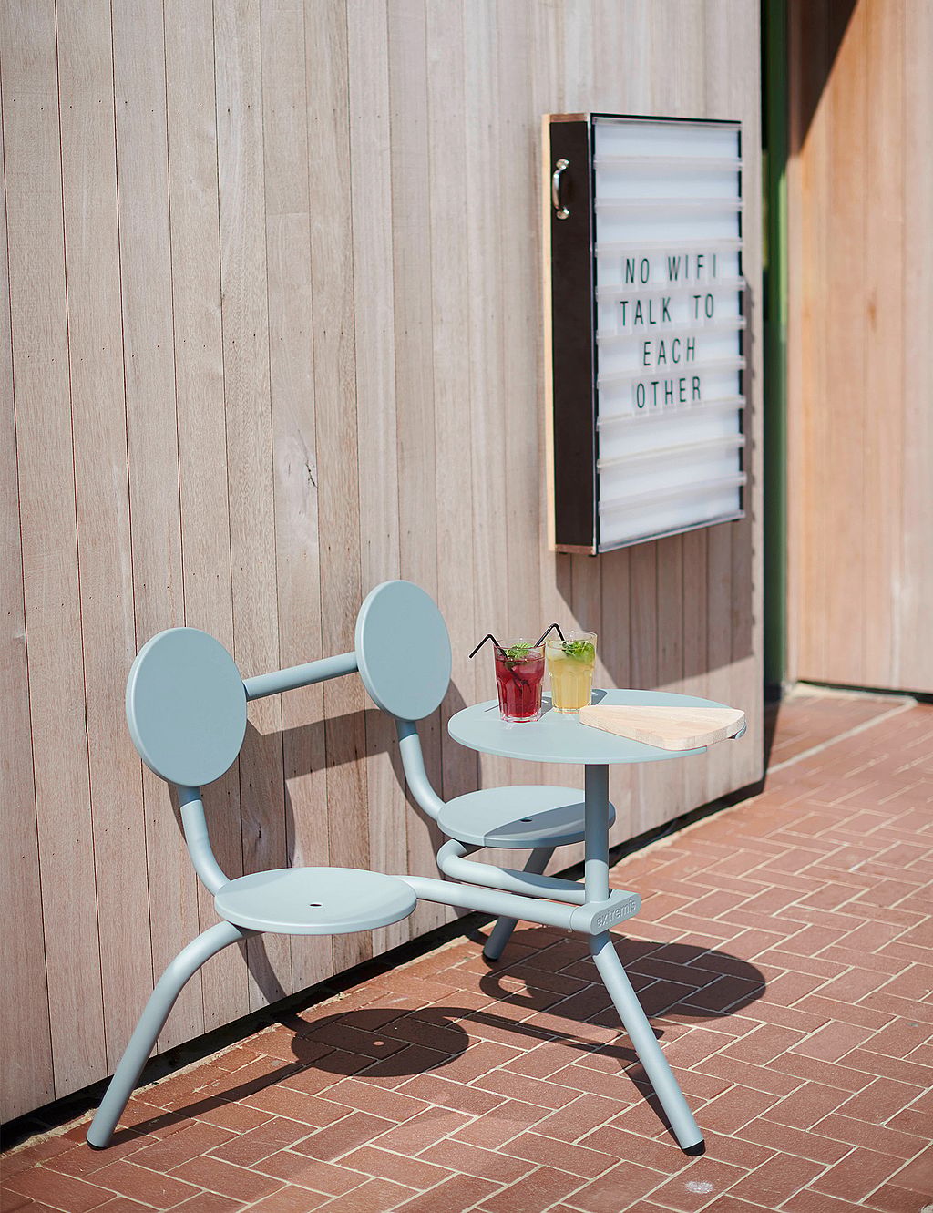 Side view of a pale blue Bistroo 2-seater with two drinks and a wooden serving board, placed beside a restaurant lightbox on a sunlit brick terrace