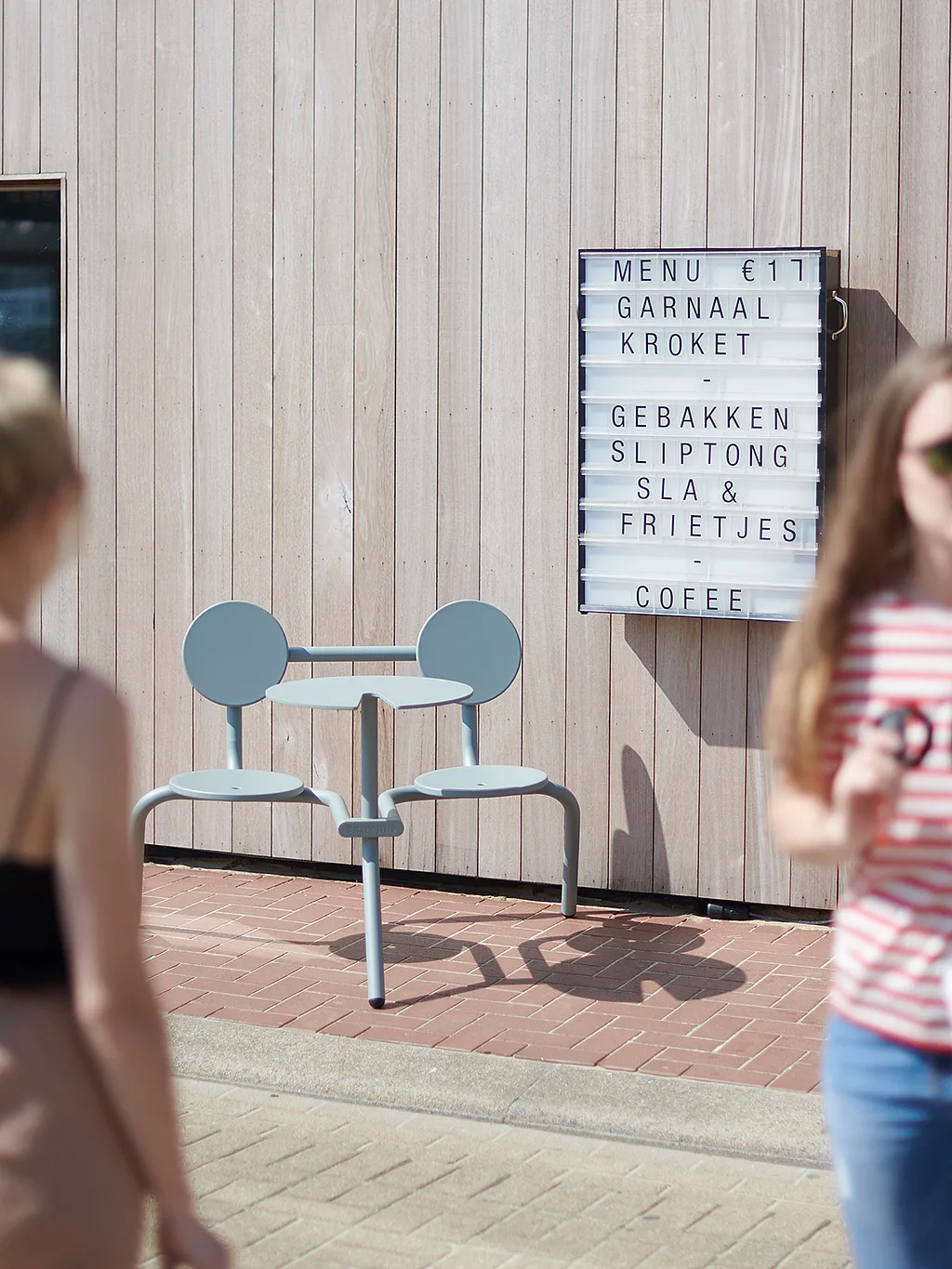 Lichtblauwe Bistroo 2-zits geplaatst op een bakstenen bestrating voor verticale houten bekleding, naast een restaurant lightbox menu, met twee voorbijgangers zacht wazig op de voorgrond.