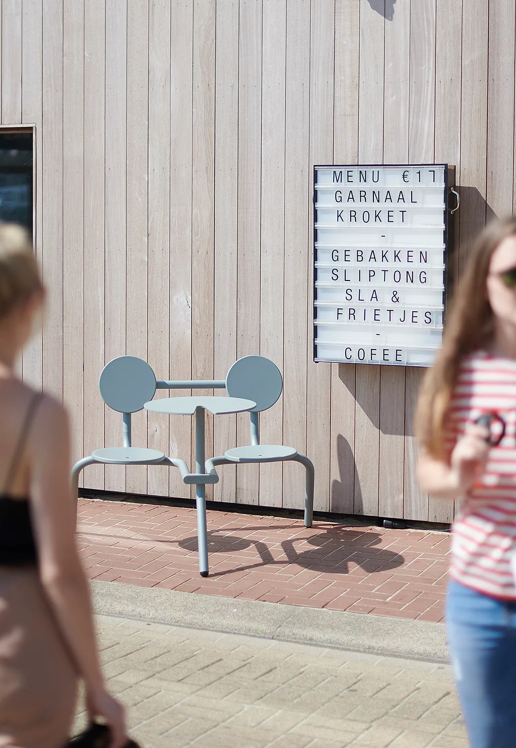 Pale blue Bistroo 2-seater placed on a brick pavement in front of vertical wooden cladding, beside a restaurant lightbox menu, with two passers-by softly blurred in the foreground.
