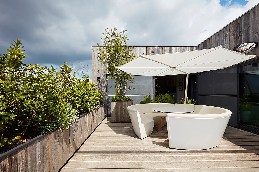 Compact dakterras met een witte Kosmos ronde bank en tafel onder een grote witte parasol, omgeven door weelderige plantbedden en verweerde houten bekleding; een gemberkat zit onder de tafel.