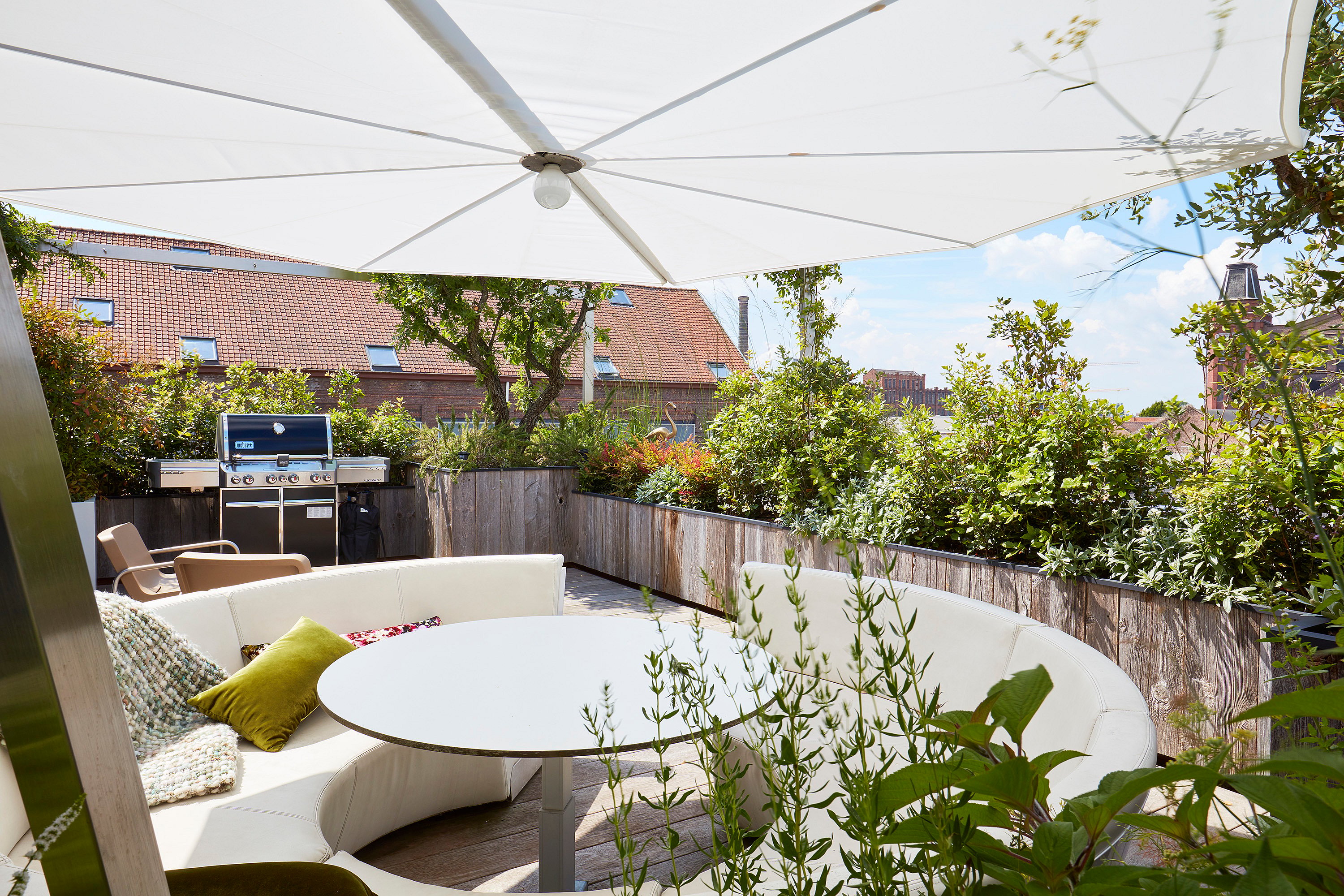 Weißer Sonnenschirm, der eine geschwungene Kosmos-Sitzanordnung auf einer sonnigen Dachterrasse beschattet, mit bepflanzten Rändern, einem Grill und Stadtdächern dahinter.