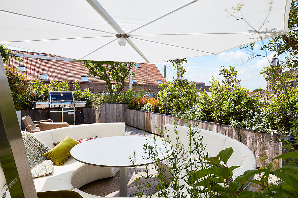 Weißer Sonnenschirm, der eine geschwungene Kosmos-Sitzanordnung auf einer sonnigen Dachterrasse beschattet, mit bepflanzten Rändern, einem Grill und Stadtdächern dahinter.