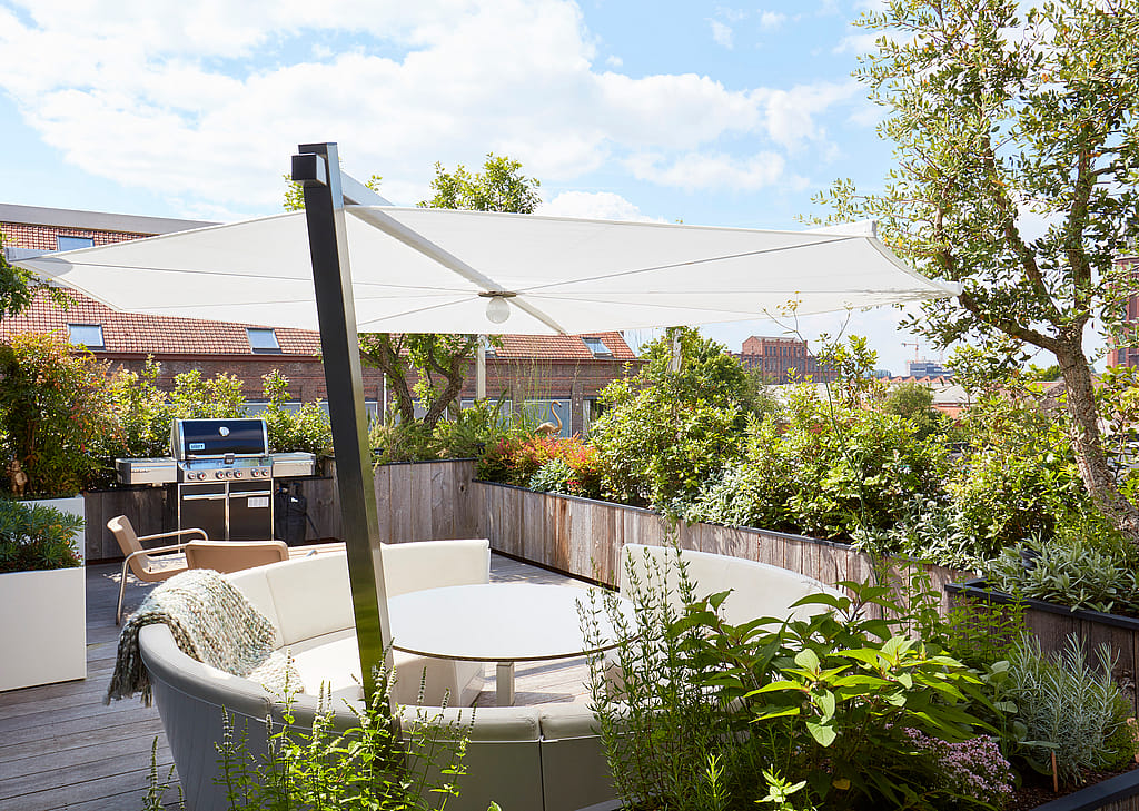 Weite Aussicht auf die bepflanzte Dachterrasse mit dem weißen Kosmos-Sofa und -Tisch unter einem großen Sonnenschirm, umgeben von Grün und mit Blick auf nahegelegene Dächer.
