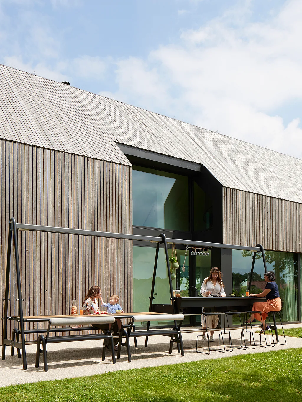 Modern timber-clad house with an AMAi outdoor table setup on the patio, where two adults and a child gather around the table and bar in the sunny garden.