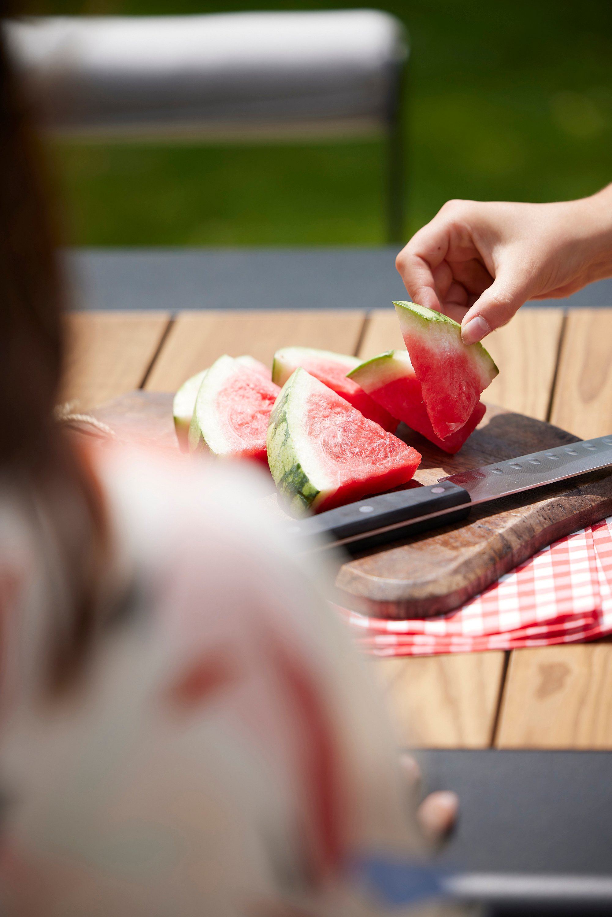   Une main soulève une tranche de pastèque d'une planche à découper posée sur la table en bois, avec un couteau et une serviette à carreaux rouges à côté.