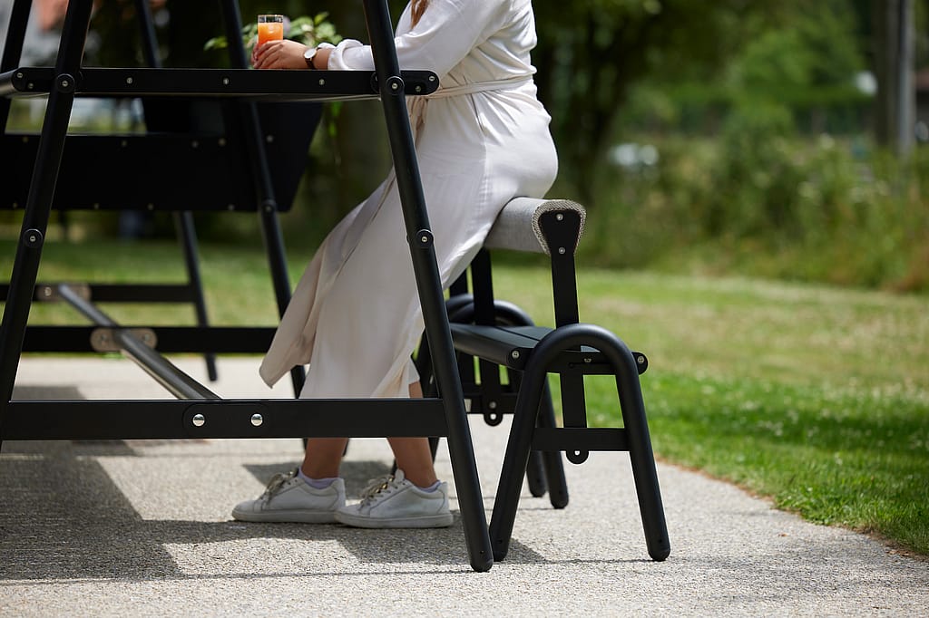 Gros plan sur une personne assise sur un tabouret haut à la table AMAi, un verre à la main, près du cadre noir, sur une terrasse ensoleillée.