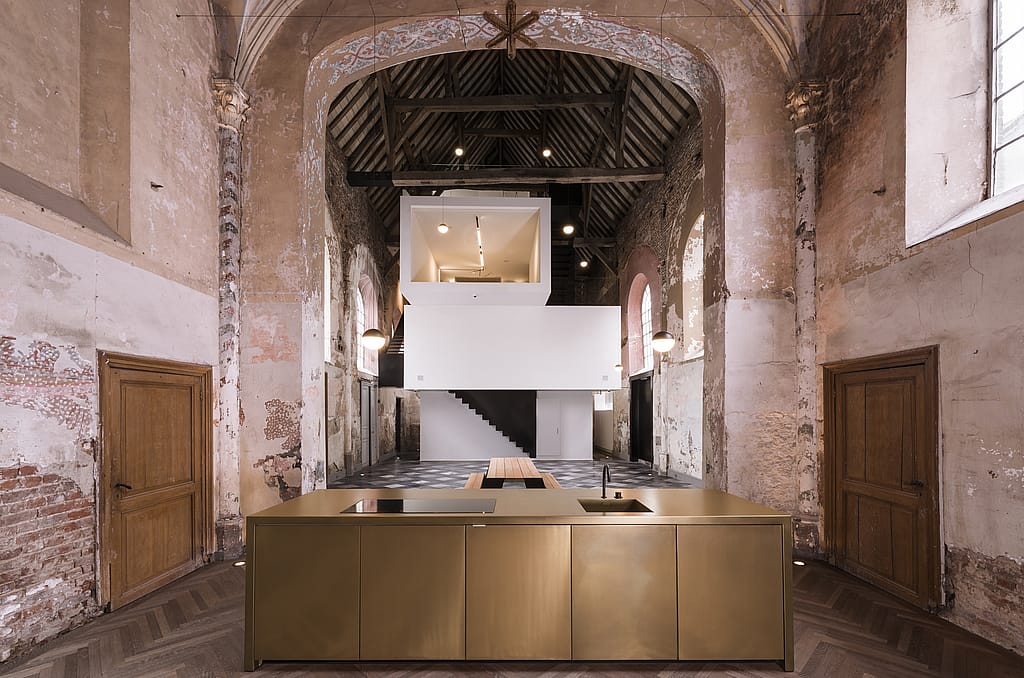 Intérieur d’une chapelle rénovée du XVIe siècle avec des murs patinés, des fermes de toit en bois apparentes, des bureaux blancs modernes et un escalier inséré au centre, un îlot de cuisine en bronze au premier plan et une table de pique-nique Hopper placée sur le sol en pierre au-delà.