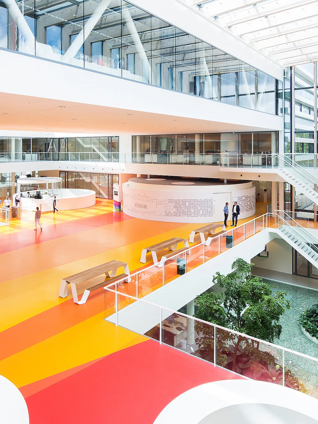 Helles mehrstöckiges Atrium bei DSM mit kräftigen gelben, orangefarbenen und roten Bodenbändern, mehreren langen Hopper-Picknicktischen entlang des Balkons und weißen Treppen, die die offenen Büroebenen verbinden.
