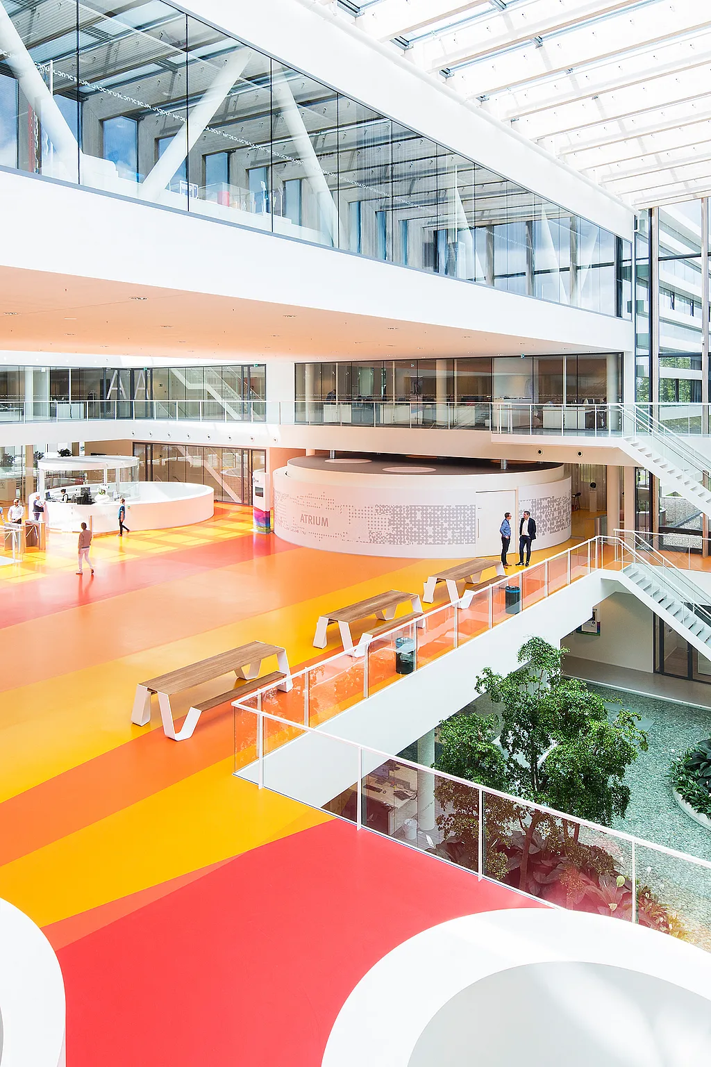 Bright multi-level atrium at DSM with bold yellow, orange, and red floor bands, several long Hopper picnic tables along the balcony, and white staircases connecting the open office levels.
