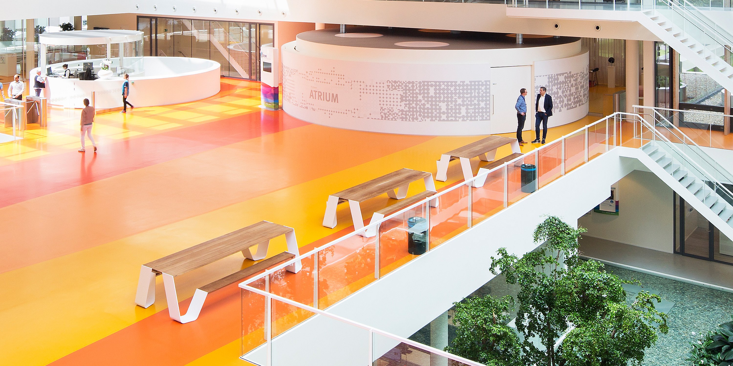 Weite Innenansicht des offenen Atriums von DSM mit kreisförmigen bepflanzten Bereichen, weißen Treppen, leuchtend orangefarbenen und rosa Gehwegen und Hopper-Tischen, die über dem sozialen Zentrum platziert sind.