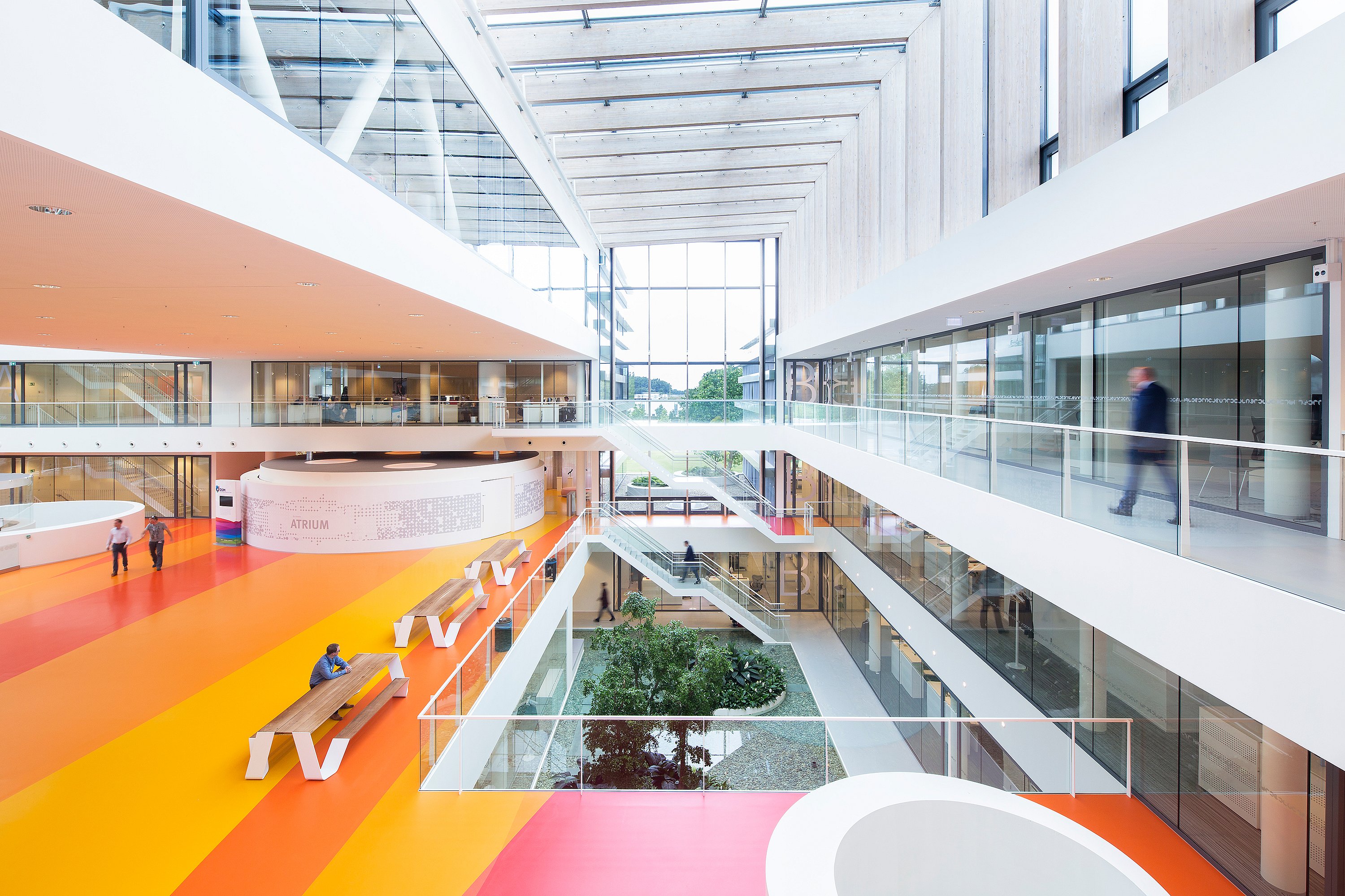 Geräumiges Atrium mit Glasdach im Forschungs- und Entwicklungszentrum von DSM mit bunt gestreiften Böden, Glasbalustraden, Innenbepflanzung darunter und Hopper-Picknicktischen für informelle Treffen