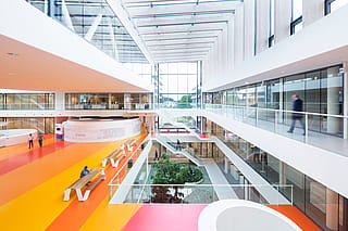 Geräumiges Atrium mit Glasdach im Forschungs- und Entwicklungszentrum von DSM mit bunt gestreiften Böden, Glasbalustraden, Innenbepflanzung darunter und Hopper-Picknicktischen für informelle Treffen