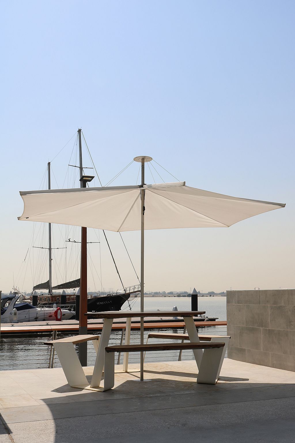 Picknicktisch White Anker, beschattet von einem Sonnenschirm von Inumbrina neben dem Yachthafen, mit Segelbootmasten und festgemachten Booten im Hintergrund.