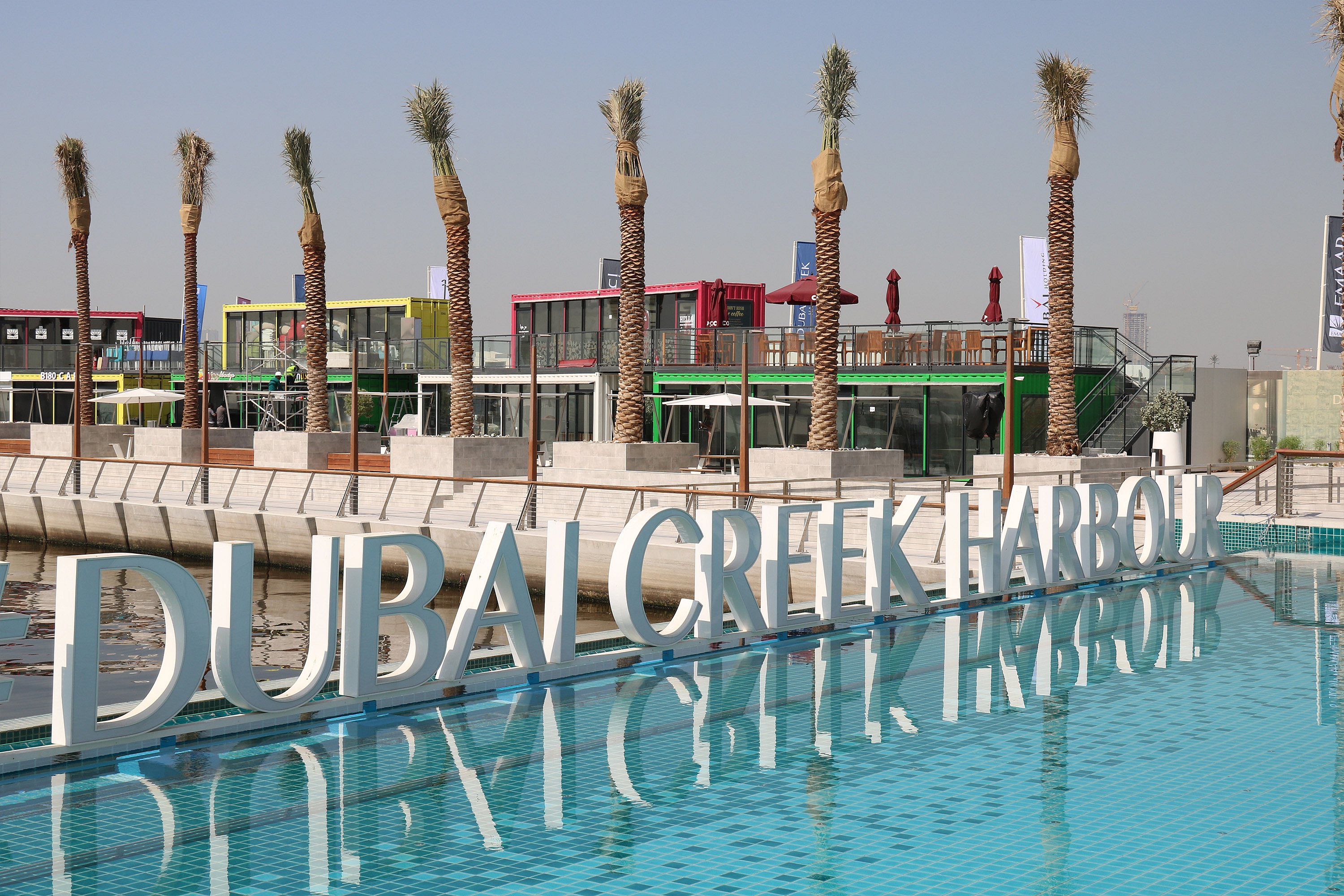 Große Buchstaben ‘im Hafen von Dubai Creek’ spiegeln sich in einem türkisfarbenen Teich, dahinter liegen Reihen von Palmen und farbenfrohe Gebäude am Wasser.