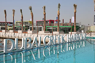 Große Buchstaben ‘im Hafen von Dubai Creek’ spiegeln sich in einem türkisfarbenen Teich, dahinter liegen Reihen von Palmen und farbenfrohe Gebäude am Wasser.