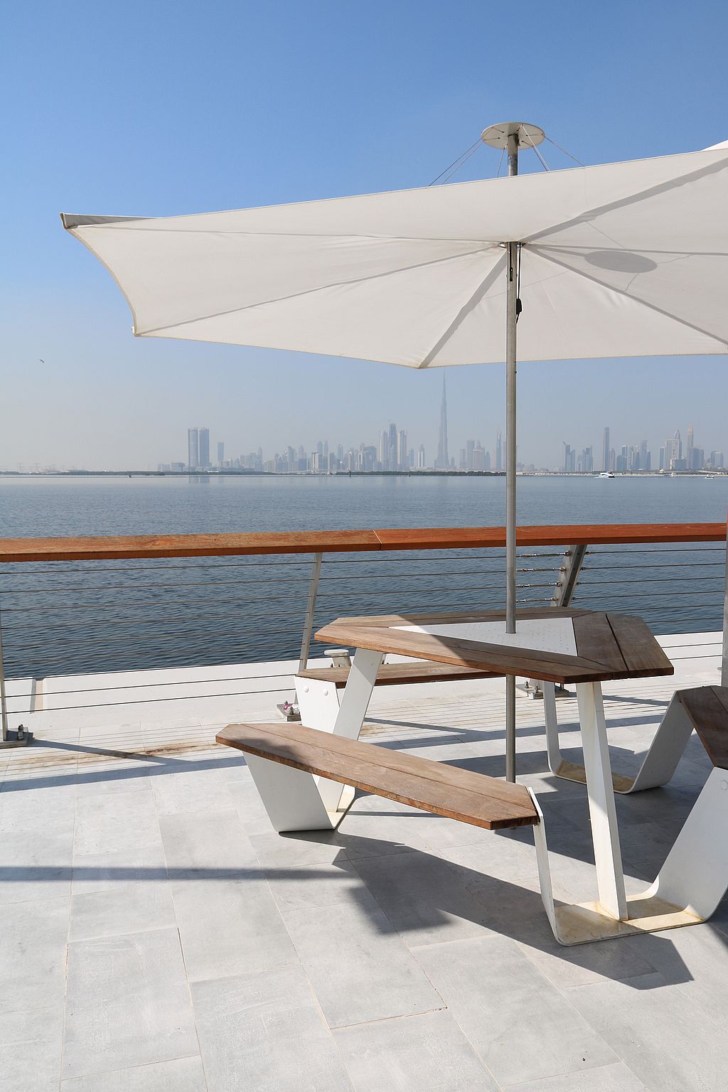 Anker-Picknicktisch mit Holzbänken unter einem großen weißen Inumbrina-Schatten auf einer Terrasse am Wasser, mit der Skyline von Dubai auf der anderen Seite des Wassers.