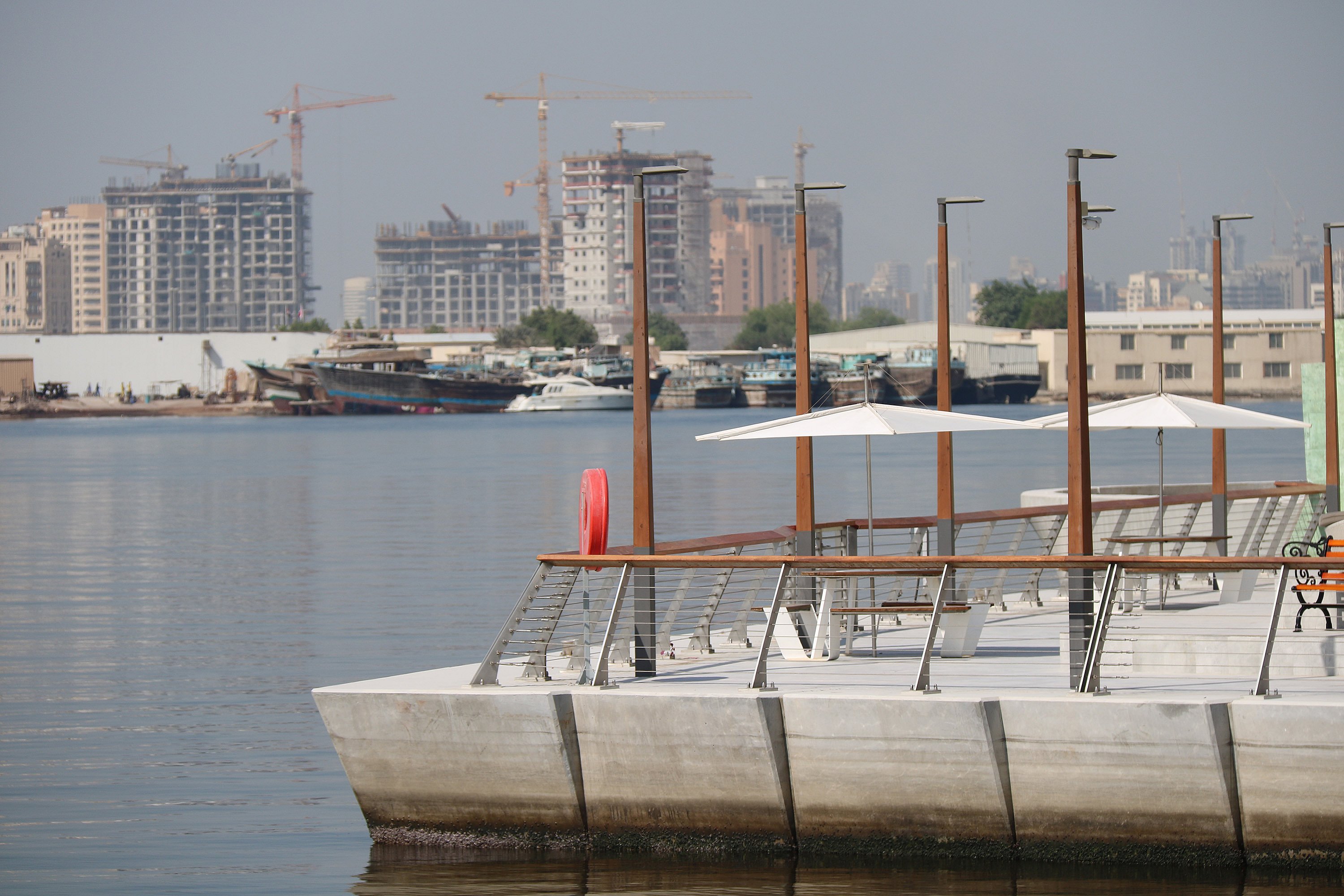 Waterfront promenade at Dubai Creek Harbour with white outdoor tables and parasols along the quay, overlooking calm water and a hazy skyline of construction cranes and buildings across the creek.