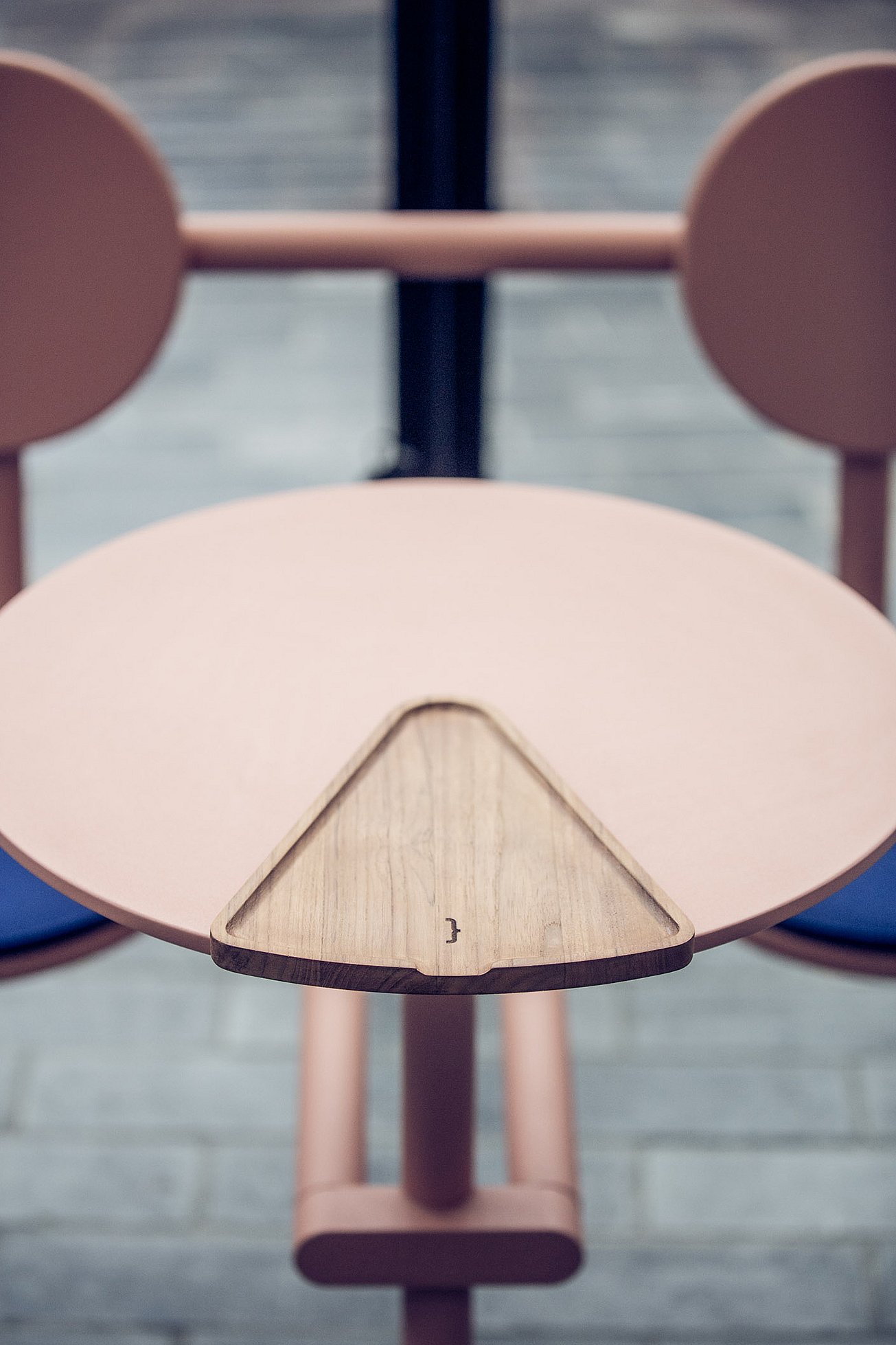Close-up van het ronde roze Bistroo tafelblad met een driehoekige houten serveerschaal netjes in het midden uitgesneden tussen de twee stoelen.