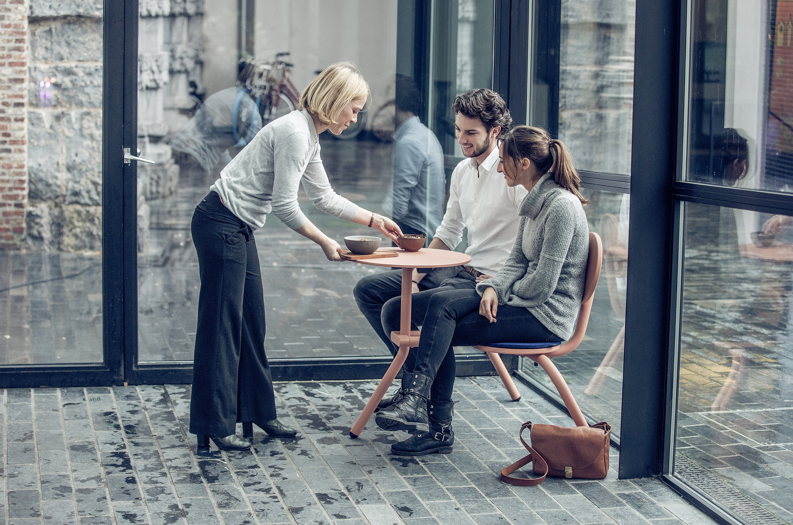 Server placing two bowls on a soft pink Bistroo table where a young couple sits side by side in a glass-walled courtyard with wet stone paving.