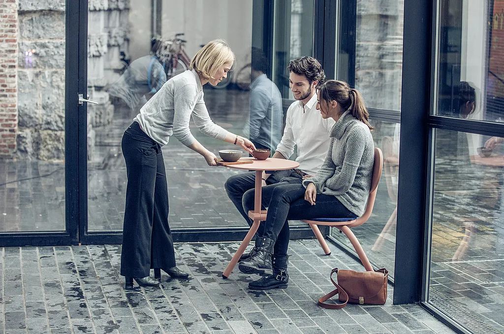 Server placing two bowls on a soft pink Bistroo table where a young couple sits side by side in a glass-walled courtyard with wet stone paving.