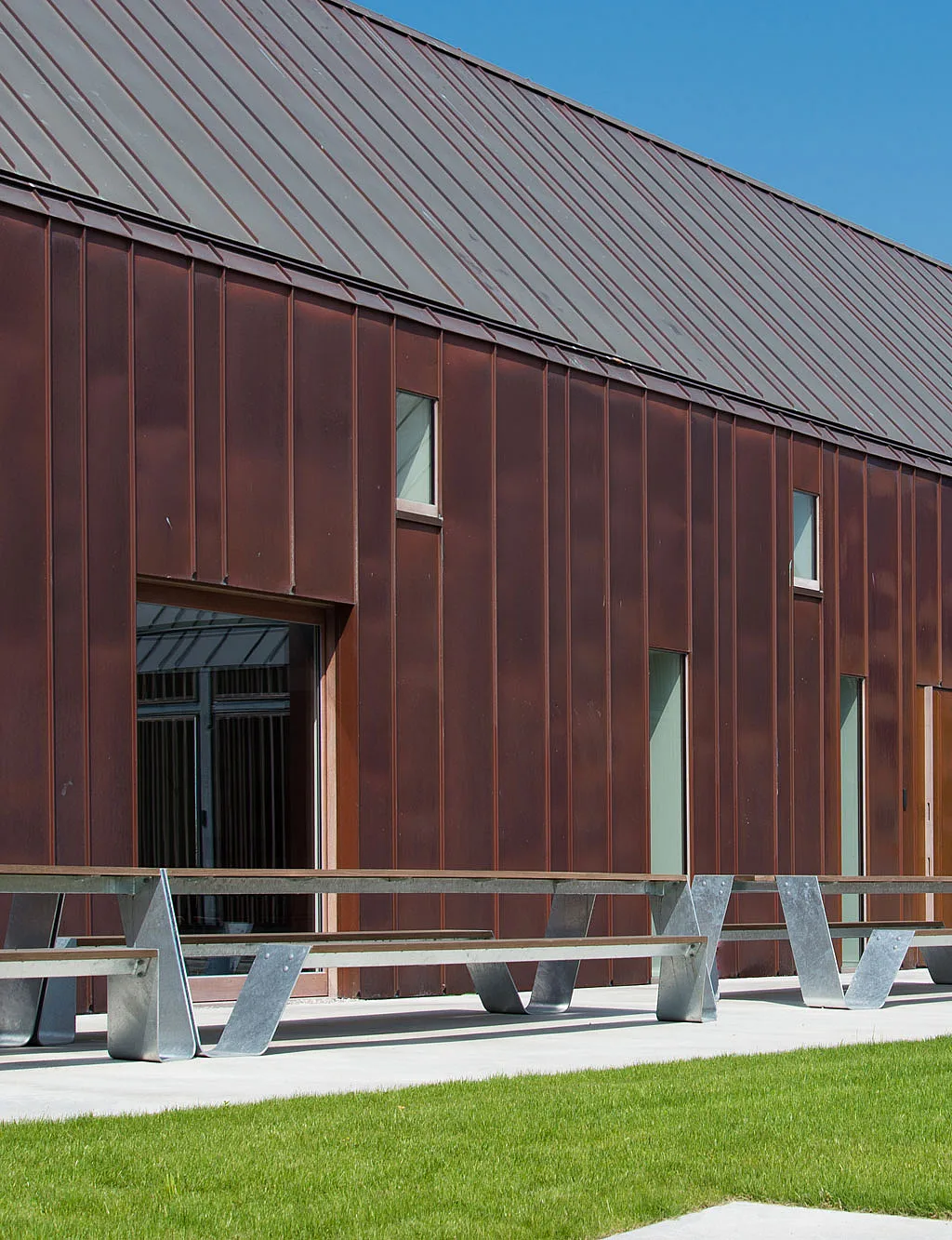 Long copper-clad holiday lodge with narrow vertical windows, fronted by several Hopper picnic tables on a paved terrace and a strip of bright green lawn.