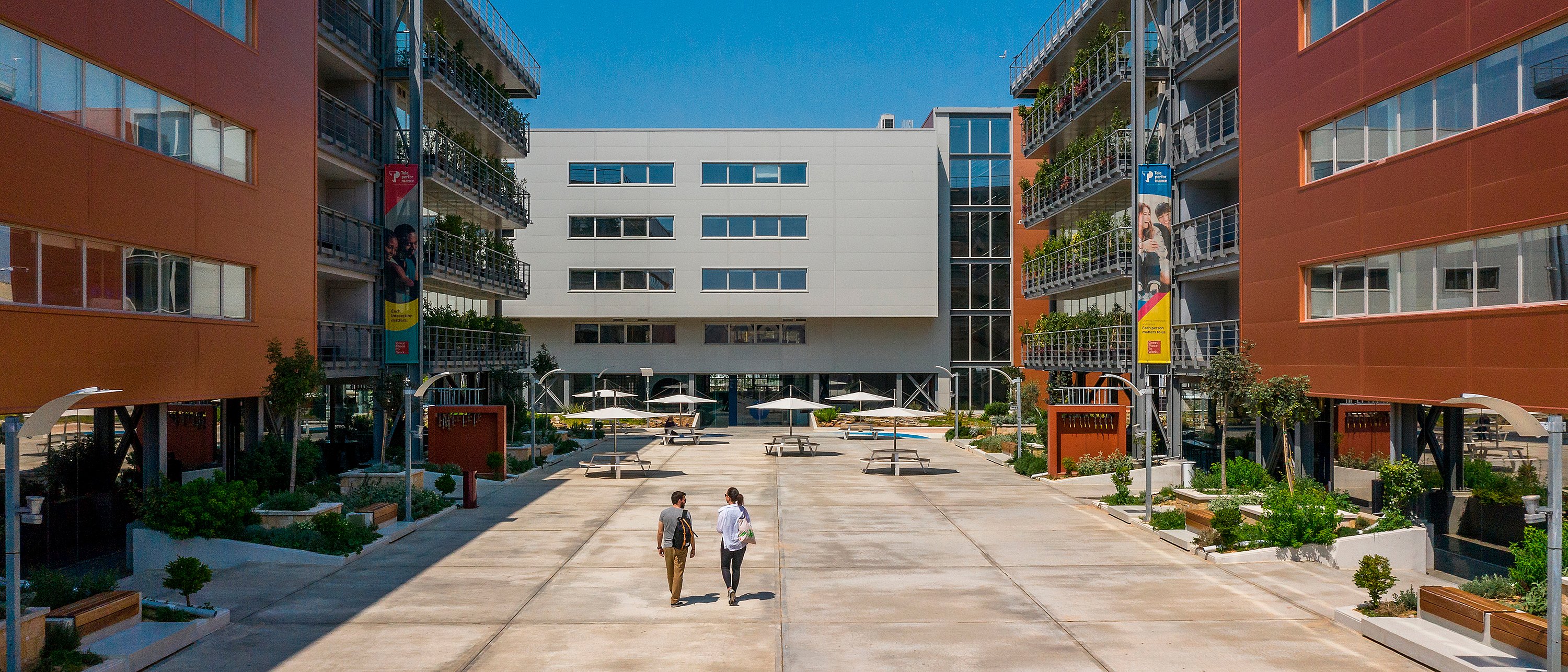Large vue sur la cour du campus de l’ENA encadrée par des immeubles de bureaux rouille et blanc, avec de larges allées, des bords plantés et plusieurs tables de pique-nique rondes ombragées réparties dans l’espace ouvert.
