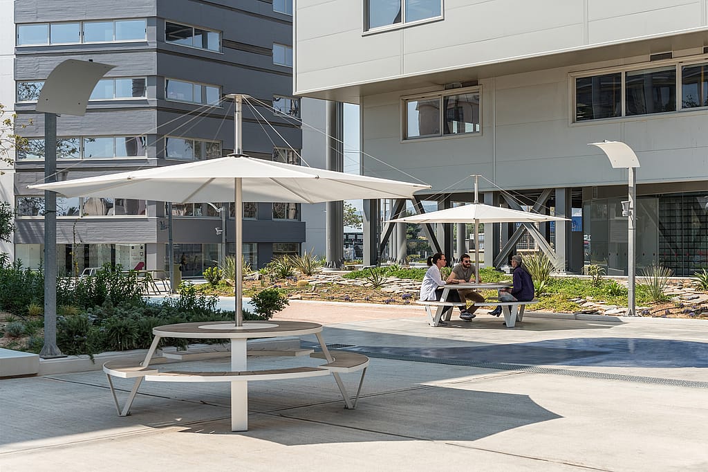 Deux tables rondes Pantagruel avec des stores blancs Inumbra se tiennent dans une cour paysagée entre des immeubles de bureaux modernes, tandis que trois collègues s’assoient ensemble à l’une des tables.