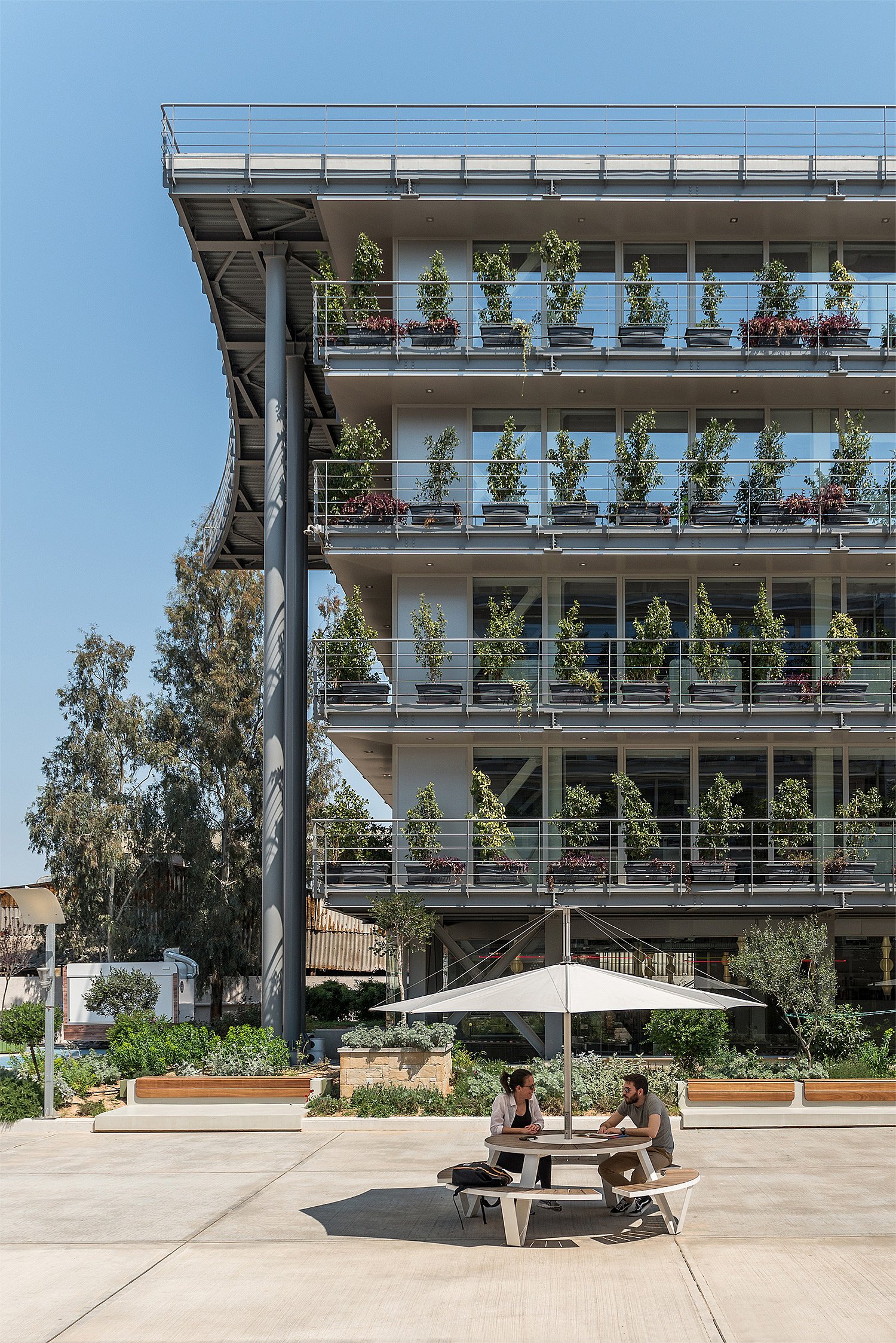Table ronde Pantagruel sous une teinte blanche Inumbra devant un bâtiment aux balcons végétaux, où deux collègues discutent au milieu de la cour pavée.