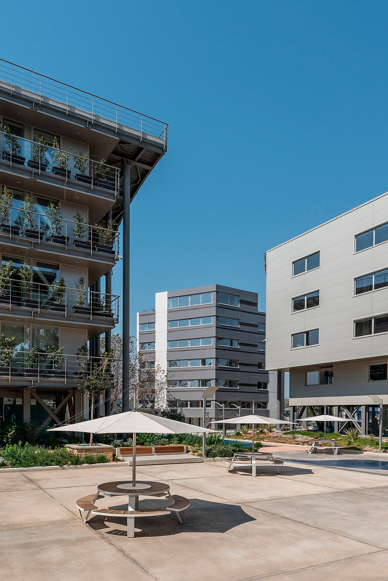 Plusieurs tables Pantagruel ombragées disposées sur une large place de bureau entre des bâtiments modernes, avec des plates-bandes basses et de fortes ombres de midi.