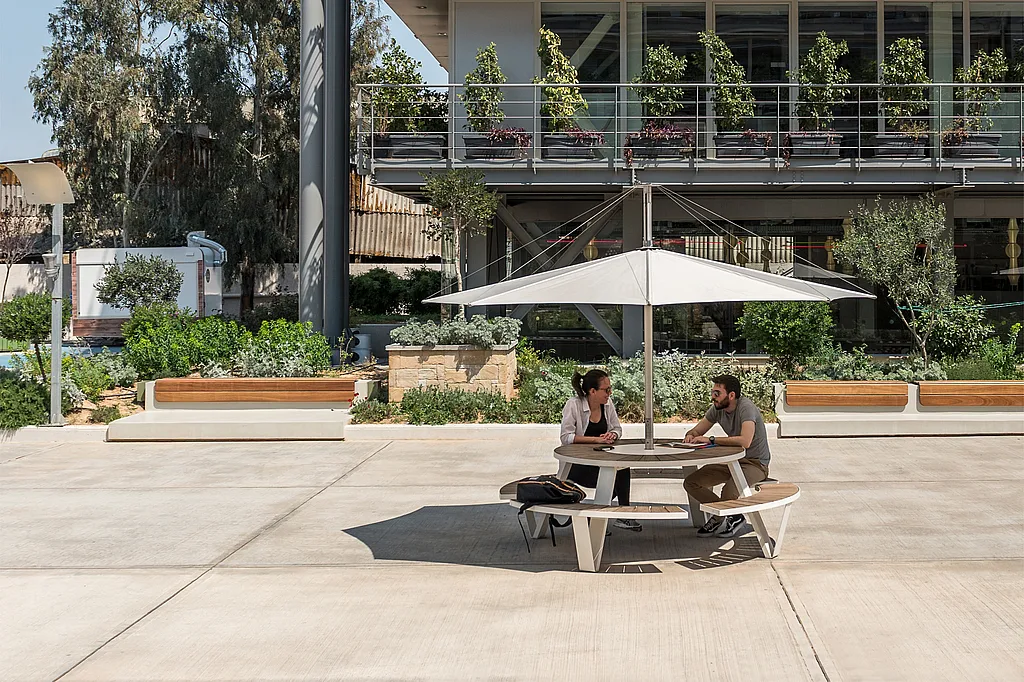 Deux collègues sont assis en face l’un de l’autre à une table de pique-nique ronde Pantagruel sous une teinte Inumbra blanche dans une spacieuse cour ensoleillée, avec des balcons plantés et un immeuble de bureaux moderne derrière eux.