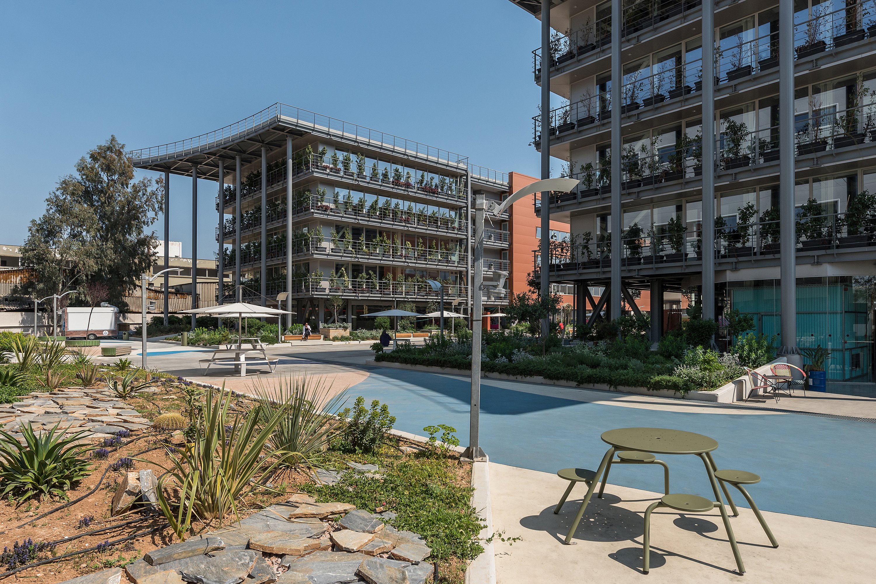 Grande cour ouverte avec des plates-bandes luxuriantes, des motifs en béton bleu et sable courbés, et plusieurs tables de pique-nique rondes sous des ombres blanches, entourées de blocs de bureaux contemporains