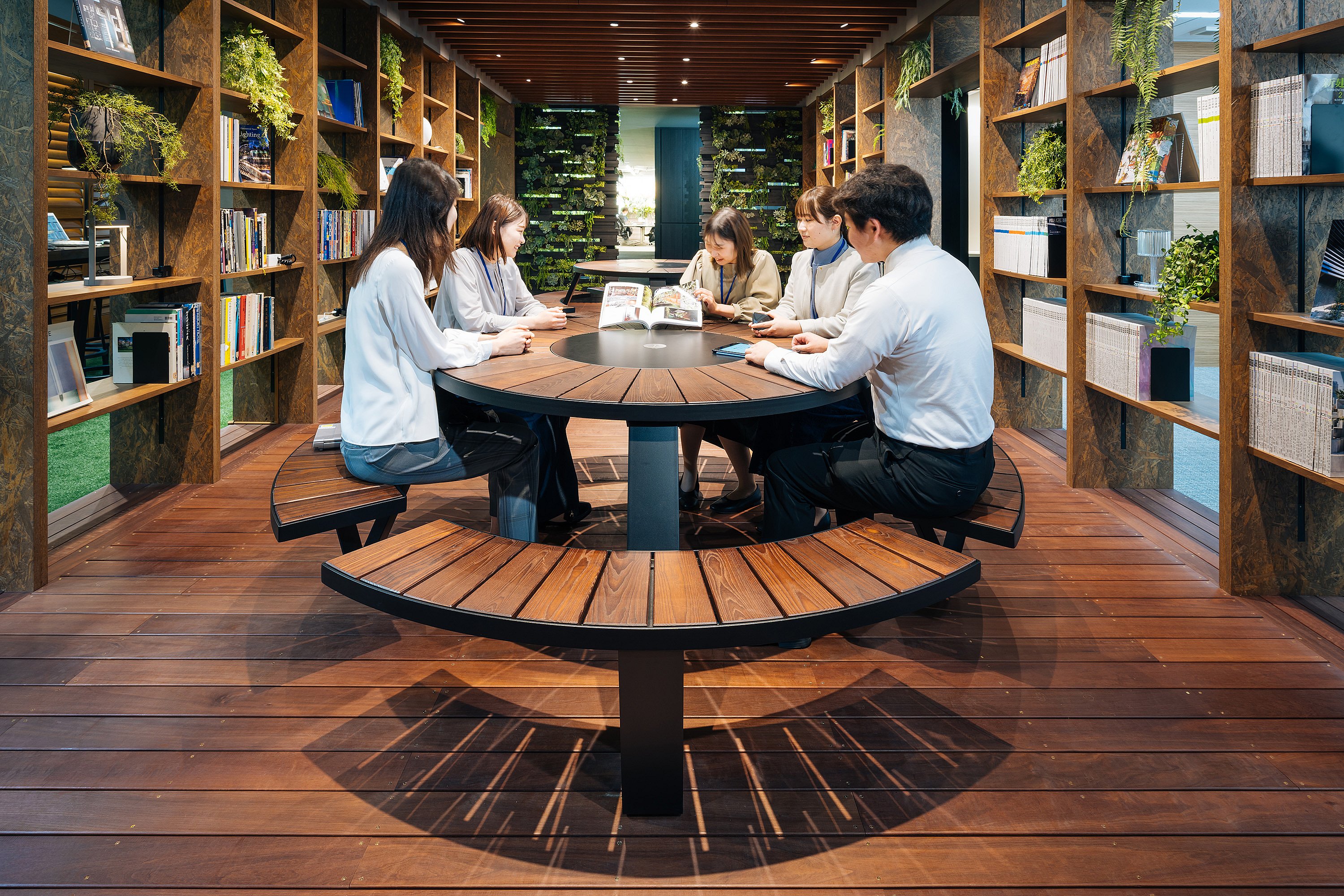 Üppiger biophiler Arbeitsbereich mit einem Pantagruel-Picknicktisch in der Mitte, der Grün, Holz und gedämpfte Beleuchtung kombiniert, um eine beruhigende Gemeinschaftszone zu schaffen.