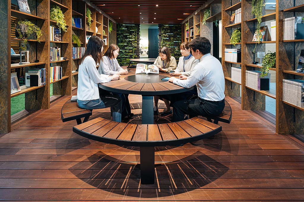 Üppiger biophiler Arbeitsbereich mit einem Pantagruel-Picknicktisch in der Mitte, der Grün, Holz und gedämpfte Beleuchtung kombiniert, um eine beruhigende Gemeinschaftszone zu schaffen.