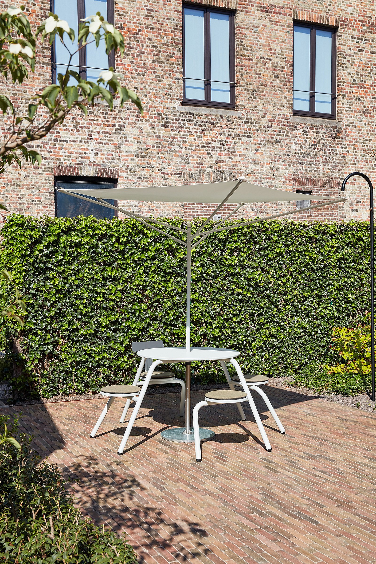   Table Virus blanc cassé avec tabourets intégrés sous un parasol en acacia clair, sur un dallage en briques rouges chaudes, devant un mur recouvert de lierre et une façade en briques historiques.