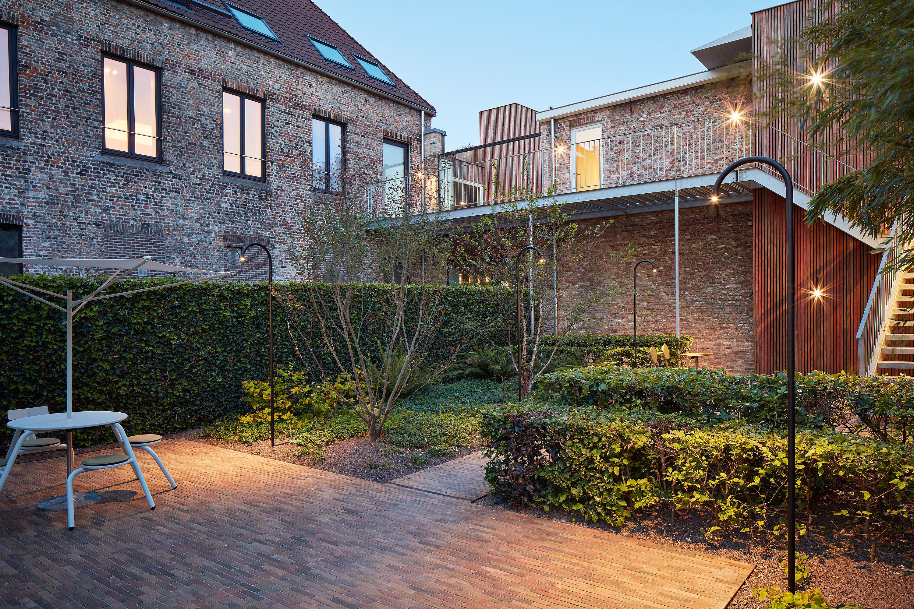   Vue au crépuscule du jardin urbain partagé, encadré par des bâtiments en briques rénovés, de jeunes arbres, des plantations basses, des luminaires de jardin noirs incurvés et le bord d'une table Virus avec parasol à gauche.