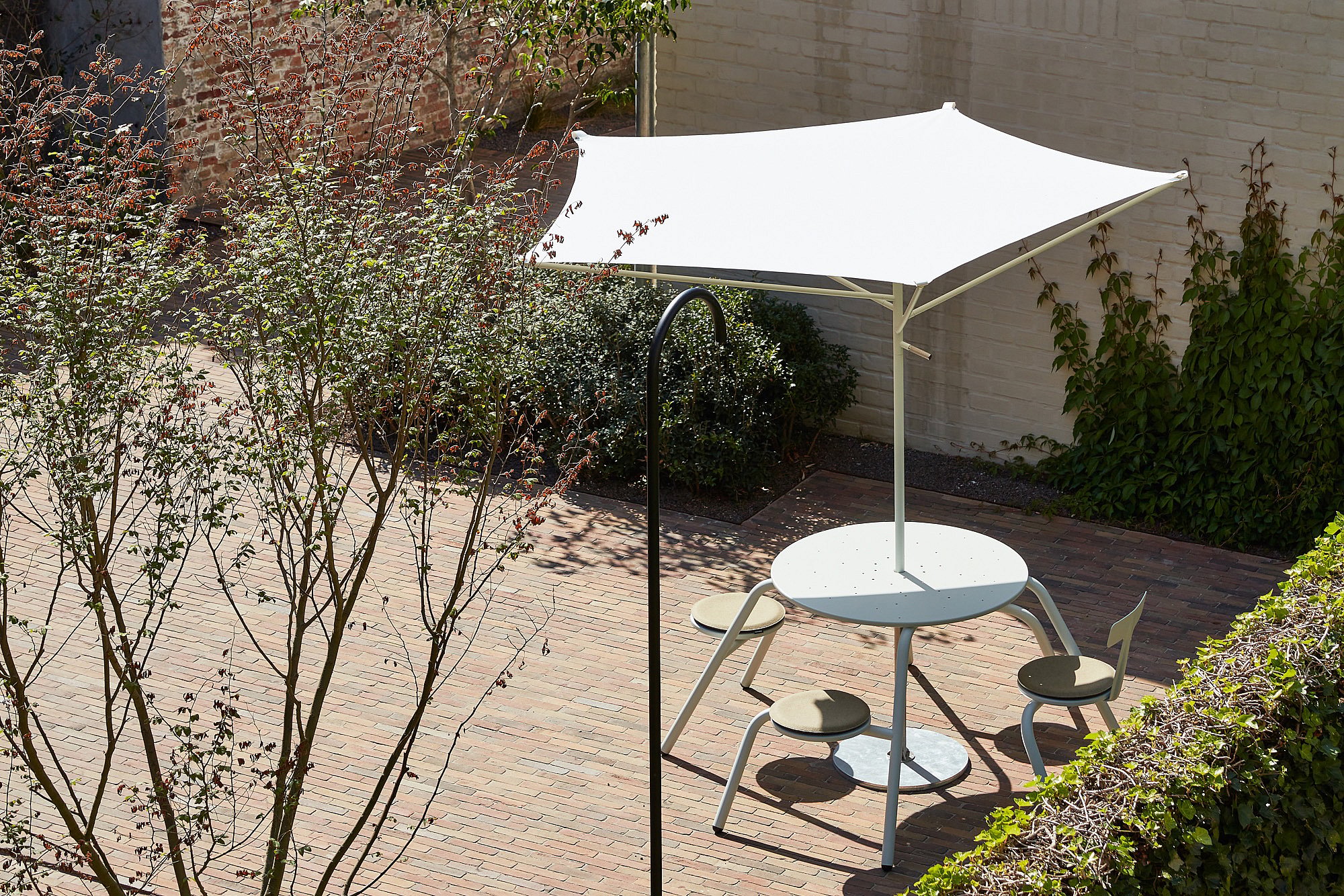 Vogelperspectief van een gedeelde bakstenen binnentuin met een crèmekleurige ronde Virus-tafel onder een witte acacia-parasol, omgeven door struiken, hagen en witgeschilderde muren.