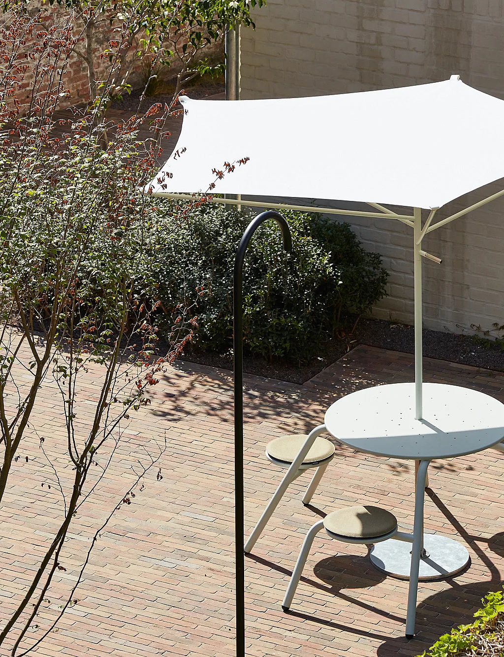 Vogelperspectief van een gedeelde bakstenen binnentuin met een crèmekleurige ronde Virus-tafel onder een witte acacia-parasol, omgeven door struiken, hagen en witgeschilderde muren.